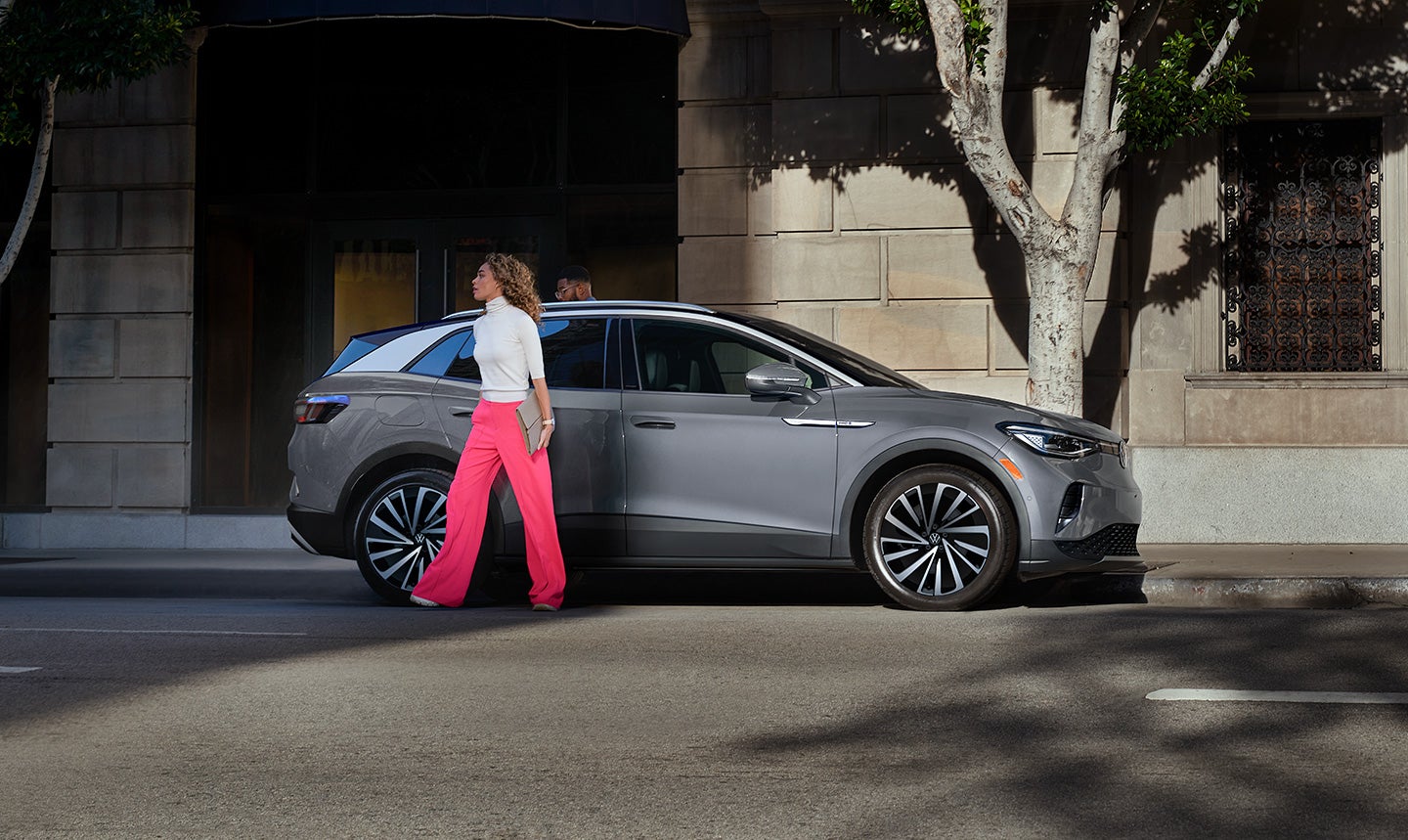 A fashionable woman walks away from the passenger side of an ID.4 Pro S Plus shown in Pure Gray, parked on a shady urban street.