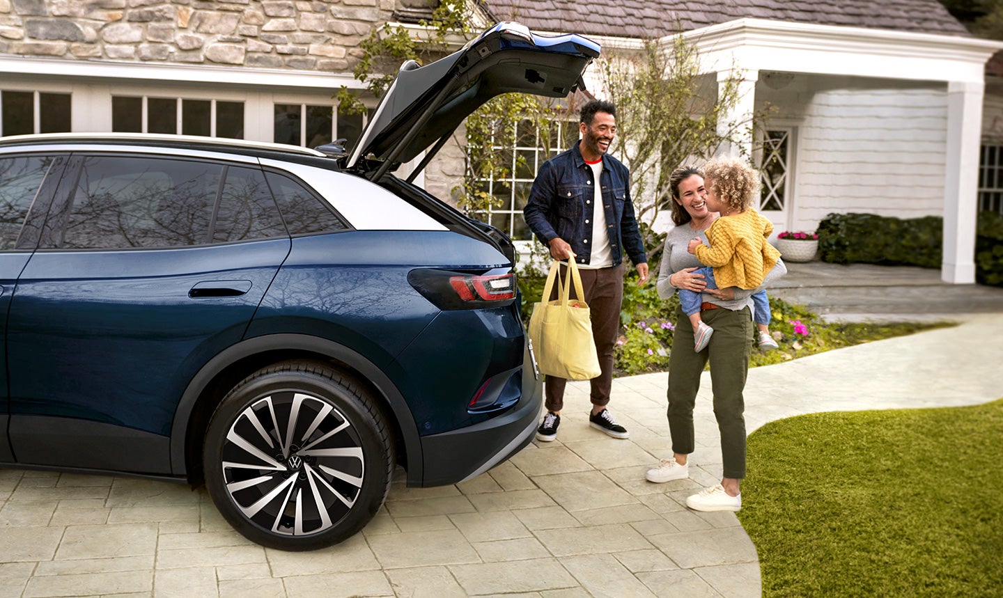 A young family loads a bag into the cargo area of an ID.4 AWD Pro S Plus shown in Tourmaline Blue Metallic parked in a residential home’s driveway.